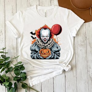 Puede incluir: Camiseta blanca con un gr&aacute;fico colorido de un payaso aterrador sosteniendo una calabaza tallada y un globo rojo. El payaso tiene el pelo rojo y una cara blanca con detalles en rojo. Murci&eacute;lagos negros en el fondo. Ropa tem&aacute;tica de Halloween.