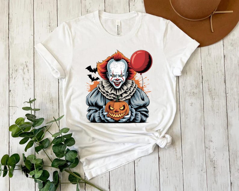 Puede incluir: Camiseta blanca con un gr&aacute;fico colorido de un payaso aterrador sosteniendo una calabaza tallada y un globo rojo. El payaso tiene el pelo rojo y una cara blanca con detalles en rojo. Murci&eacute;lagos negros en el fondo. Ropa tem&aacute;tica de Halloween.