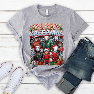Puede incluir: Camiseta gris claro con la frase "MERRY CREEPMAS" e ilustraciones de personajes de pel&iacute;culas de terror con gorros de Pap&aacute; Noel. La camiseta se muestra con vaqueros azules y zapatillas blancas.