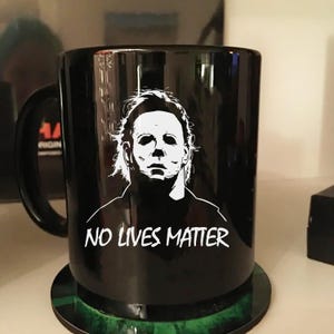 以下が含まれることがあります： 白のホラー映画のキャラクターのグラフィックと「NO LIVES MATTER」の文字が入った黒いセラミックマグ。マグには黒いハンドルが付いており、緑と黒のコースターの上に置かれています。ホラー映画ファンへのギフトに最適です。