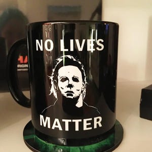 以下が含まれることがあります： 「NO LIVES MATTER」の文字とキャラクターの白いグラフィックが描かれた黒いセラミックマグカップ。マグカップには黒いハンドルがあり、緑と黒のコースターの上に置かれています。ホラーをテーマにしたアイテムです。