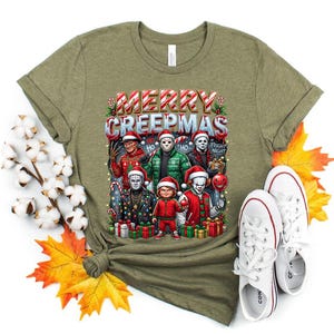 Puede incluir: Camiseta verde oliva con un gr&aacute;fico festivo de personajes de pel&iacute;culas de terror con gorros de Pap&aacute; Noel y el texto "MERRY CREEPMAS". El dise&ntilde;o incluye regalos envueltos y luces navide&ntilde;as. Zapatillas blancas junto a la camiseta.