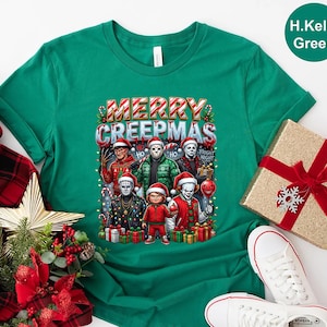 Puede incluir: Una camiseta verde con el texto "MERRY CREEPMAS" e im&aacute;genes de personajes de pel&iacute;culas de terror con gorros de Pap&aacute; Noel. La camiseta tiene cuello redondo y mangas cortas. Una caja de regalo con un lazo rojo est&aacute; en el fondo.