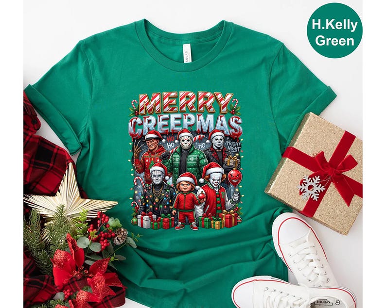 Puede incluir: Una camiseta verde con el texto "MERRY CREEPMAS" e im&aacute;genes de personajes de pel&iacute;culas de terror con gorros de Pap&aacute; Noel. La camiseta tiene cuello redondo y mangas cortas. Una caja de regalo con un lazo rojo est&aacute; en el fondo.