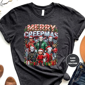 Puede incluir: Camiseta gris oscuro jaspeado con el texto "MERRY CREEPMAS" e im&aacute;genes de personajes de pel&iacute;culas de terror con gorros de Pap&aacute; Noel. El dise&ntilde;o incluye bastones de caramelo, luces navide&ntilde;as y cajas de regalo. La camiseta es de cuello redondo.