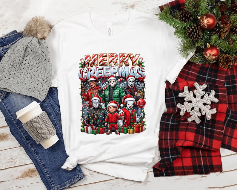 Puede incluir: Camiseta blanca con un gr&aacute;fico colorido que dice "MERRY CREEPMAS" con personajes de pel&iacute;culas de terror con gorros de Pap&aacute; Noel. El dise&ntilde;o incluye bastones de caramelo y regalos. La camiseta se muestra con un gorro gris, jeans y una bufanda a cuadros roja y negra.