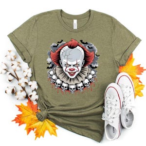 Puede incluir: Camiseta verde oliva con un gr&aacute;fico de payaso aterrador. El payaso tiene pelo rojo, cara blanca y un cuello con volantes adornado con calaveras. El dise&ntilde;o incluye murci&eacute;lagos y una cabeza agrietada. Zapatillas blancas con detalles rojos en la parte inferior.