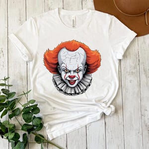 Puede incluir: Camiseta blanca con un gr&aacute;fico de un payaso aterrador con pelo rojo, cara blanca y cuello con volantes. El payaso tiene una expresi&oacute;n siniestra. La camiseta est&aacute; hecha de un material suave.