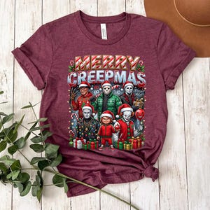 Puede incluir: Camiseta granate con el texto "MERRY CREEPMAS" sobre un gr&aacute;fico de personajes de pel&iacute;culas de terror con gorros de Pap&aacute; Noel. El gr&aacute;fico incluye personajes como Freddy Krueger y Jason Voorhees, rodeados de decoraciones navide&ntilde;as.