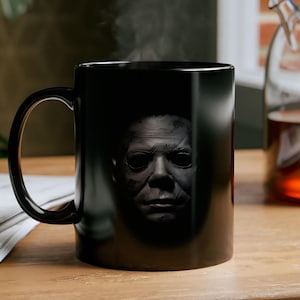 Op de afbeelding: Zwarte keramische mok met een grijswaardenafbeelding van een menselijk gezicht. De mok heeft een gebogen handvat en bevat een stomende drank. De mok staat op een houten oppervlak. De mok is ongeveer 10 cm hoog.