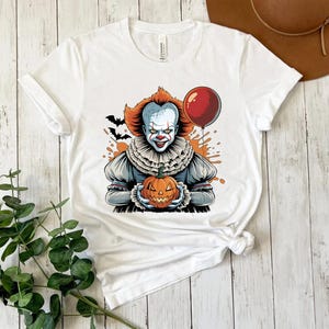 Puede incluir: Camiseta blanca con un dise&ntilde;o de Halloween. El dise&ntilde;o presenta un payaso amenazante sosteniendo una calabaza tallada y un globo rojo. El payaso tiene pelo naranja y un cuello con volantes, con murci&eacute;lagos y salpicaduras naranjas en el fondo.