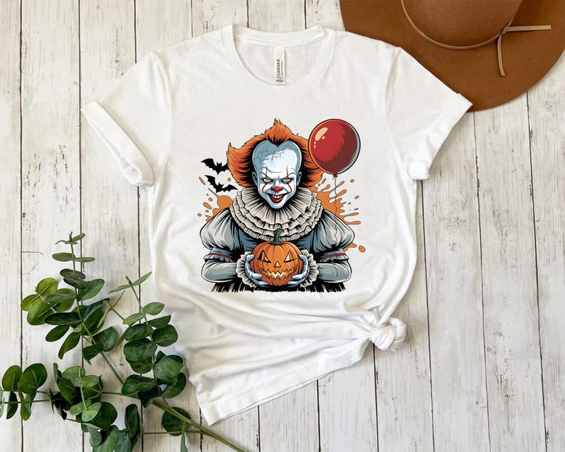 Puede incluir: Camiseta blanca con un dise&ntilde;o de Halloween. El dise&ntilde;o presenta un payaso amenazante sosteniendo una calabaza tallada y un globo rojo. El payaso tiene pelo naranja y un cuello con volantes, con murci&eacute;lagos y salpicaduras naranjas en el fondo.