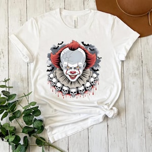 Puede incluir: Camiseta blanca con un gr&aacute;fico de un payaso aterrador con pelo rojo, cara blanca y nariz roja. El payaso est&aacute; rodeado de murci&eacute;lagos y calaveras. La camiseta es de cuello redondo y tiene mangas cortas.