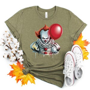 Puede incluir: Camiseta verde oliva con un gr&aacute;fico de un payaso aterrador sosteniendo un globo rojo. El payaso tiene pelo rojo, pintura facial blanca y un cuello con volantes. Zapatillas blancas con detalles rojos est&aacute;n al lado de la camiseta.
