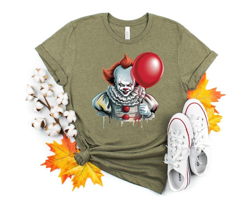 Puede incluir: Camiseta verde oliva con un gr&aacute;fico de un payaso aterrador sosteniendo un globo rojo. El payaso tiene pelo rojo, pintura facial blanca y un cuello con volantes. Zapatillas blancas con detalles rojos est&aacute;n al lado de la camiseta.