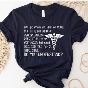 May include: Navy blue t-shirt with white text and a medical symbol. The text includes medical abbreviations and the question "DO YOU UNDERSTAND?" The shirt is folded at the bottom.