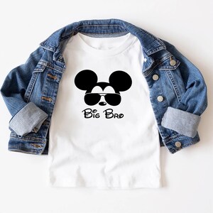 以下が含まれることがあります： サングラスをかけたミッキーマウスの黒いグラフィックと「Big Bro」の文字が入った白いTシャツ。このTシャツは、青いデニムジャケットと組み合わせています。