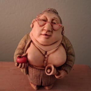 Puede incluir: Figura de una mujer con gafas rosas, camisa abotonada y falda a cuadros. Sostiene una manzana roja y un rollo de papel. La figura está pintada a mano con detalles.