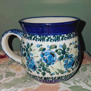May include: A ceramic mug with a rounded body and a handle. The mug features a blue floral pattern on a white background, with a dark blue rim and handle accents. The design includes blue flowers and green leaves.