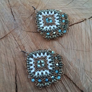 Può includere: Un paio di orecchini pendenti con un design geometrico. Gli orecchini sono realizzati con perline bianche, marroni e blu.