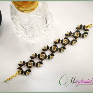 Peut inclure: Un bracelet en perles noires et dorées avec des accents verts. Le bracelet est composé de petites perles rondes enfilées ensemble dans un motif de carrés. Le bracelet a un fermoir en métal doré.