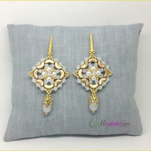 Op de afbeelding: Een paar gouden en witte kralen oorbellen met een vierkant ontwerp en een traanvormige hanger. De oorbellen worden getoond op een grijs stoffen oppervlak.