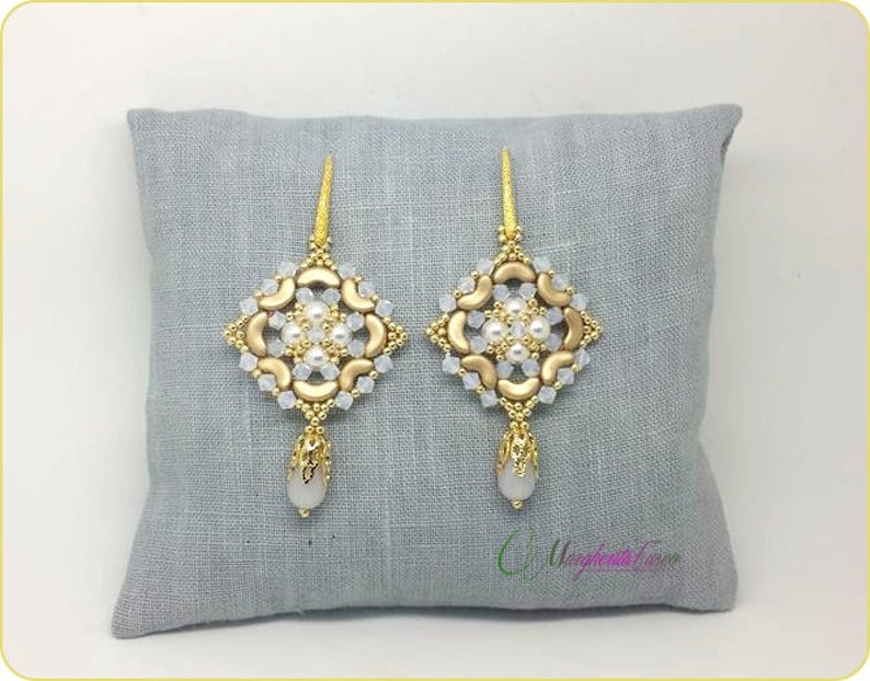 Może przedstawiać: Para złotych i białych koralikowych kolczyk&oacute;w z kwadratowym wzorem i wisiorem w kształcie łzy. Kolczyki są prezentowane na szarej powierzchni z tkaniny.