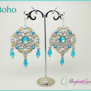 Könnte beinhalten: Silberne und goldene Perlenohrringe mit einem türkisfarbenen Kristall in der Mitte und türkisfarbenen Tropfenperlen, die unten hängen. Die Ohrringe werden auf einem silbernen Ständer präsentiert. Boho.