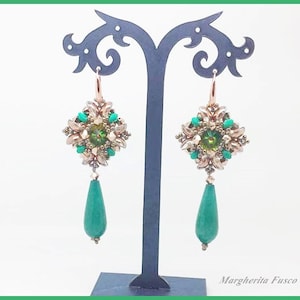 Pode incluir: Um par de brincos pendentes com um design geométrico verde e dourado. Os brincos estão pendurados em um suporte preto. Os brincos são feitos de contas e têm uma pedra verde em forma de lágrima na parte inferior.