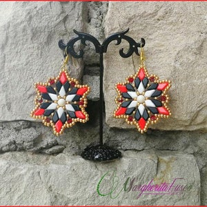 May include: A pair of star-shaped earrings made with red, black, white, and gold beads. The earrings are hanging from a black stand.