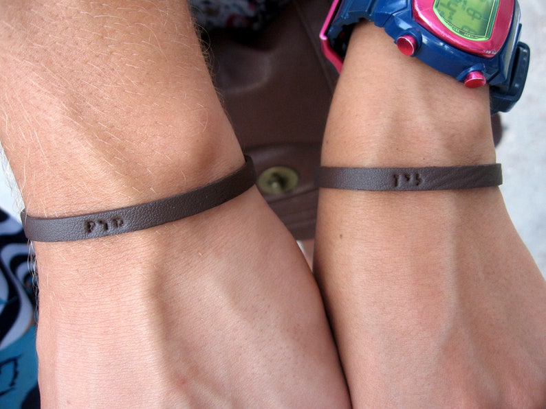 Bracelet on a man wrist made in look alike leather designed with Hebrew Name personalized on it alongside the wrist of a woman wearing the same bracelet with her name on it in hand stamped on the leather in Hebrew letters.