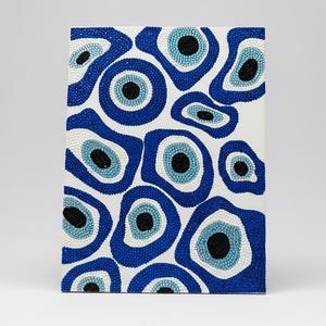 May include: A rectangular canvas with a pattern of blue and white beaded evil eye designs. The eyes vary in size and shape, with a black pupil in the centre of each eye. The background is white.