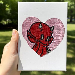 May include: A white canvas art piece featuring a red devil character with horns, wings, and a tail, set against a pink heart background. The artwork is embellished with small, sparkling details.