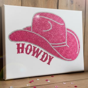 Puede incluir: Arte en lienzo con un diseño de sombrero de vaquero rosa adornado con pedrería. La palabra "HOWDY" está escrita con pedrería rosa debajo del sombrero. La obra de arte está sobre un fondo blanco.