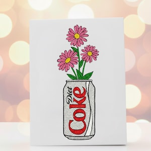 May include: A white canvas print featuring a Diet Coke can with the word "Coke" in red rhinestones. Pink flowers with yellow centers and green stems emerge from the can, creating a unique floral arrangement.