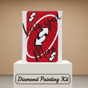 May include: A diamond painting kit featuring a red card with the words "Love You More" and white arrows. The card is displayed on a white pedestal, with the words "Diamond Painting Kit" below.