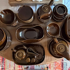 May include: A collection of dark brown ceramic dinnerware including teapots, bowls, cups, and serving trays. The set features a glossy, uniform finish across all pieces, with various rounded shapes, including oval serving dishes and lidded teapots.