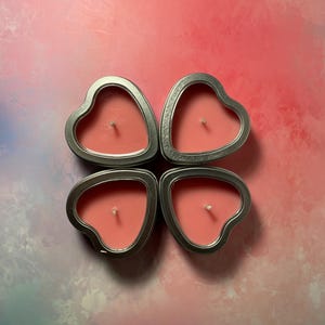 May include: Four heart-shaped candles in silver tins. The candles are a light pink color and have a single wick in the center.