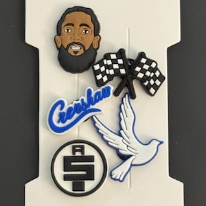 May include: Set of five shoe charms featuring a black and white checkered flag, a white dove, a black and white logo with the letter "A" and the letter "S" inside a circle, a blue and white logo with the word "Crenshaw" in a cursive font, and a cartoon image of a man with a beard and a black beanie.