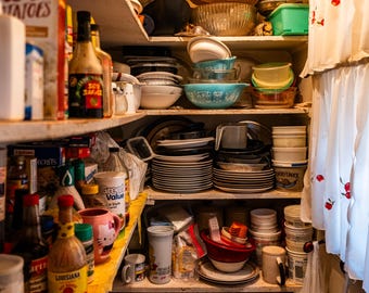 May include: A pantry filled with various kitchen items. Shelves hold stacks of plates, bowls, and containers. Bottles of sauces and jars of food are visible. A white curtain with red cherry embroidery hangs on the right.