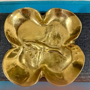 May include: A brass-colored, four-lobed dish with a bird-like figure in the center. The dish has a shiny, reflective surface and is likely made of metal. The dish is resting on a dark surface, possibly a book or a similar item.
