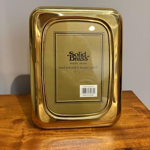 May include: A rectangular gold-colored solid brass photo frame with rounded corners. The frame features a polished, lacquer-coated finish. The insert reads Solid Brass PHOTO FRAME hand polished & lacquer coated 5" x 7" with a barcode label.