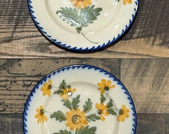Könnte beinhalten: Zwei runde Keramikteller mit cremefarbener Basis, handbemalt mit gelben Blumen und grünen Blättern in der Mitte. Die Ränder sind mit einem dunkelblauen Wellenmuster verziert. Die Teller haben eine glänzende Oberfläche und eine glatte Textur.