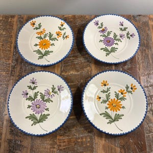 May include: A set of four round ceramic saucers with a cream-colored base and a blue scalloped edge. Each saucer features a hand-painted floral motif in the center, alternating between orange and purple daisy-like flowers with green leaves.