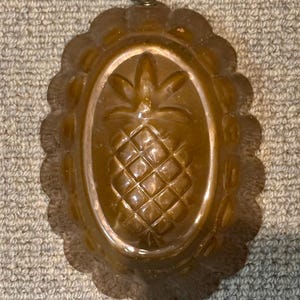 May include: A vintage, oval-shaped copper mold with a pineapple design in the center. The mold has a scalloped edge and a small wire loop for hanging. The pineapple is detailed with a diamond pattern.