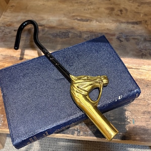 May include: A metal boot pull with a polished gold-tone horse head handle and a long, curved black shaft. The horse head features detailed facial contours, including an eye and ear, while the black shaft ends in a smooth, rounded hook.