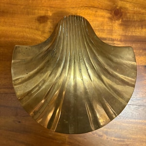May include: A polished brass dish shaped like a scallop shell. The object features a series of raised, radiating ridges that extend from the center to the scalloped outer edge, creating a fan-like appearance with a metallic gold-toned finish.