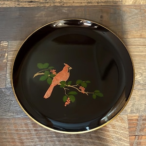 May include: A round, black lacquer tray featuring a central illustration of a red cardinal bird perched on a branch with green leaves and small red berries. The tray has a smooth, glossy finish and a thin gold-colored rim around the edge.