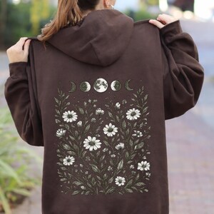 May include: Dark chocolate brown hoodie with a floral and moon phase design on the back. The graphic features white daisies, green leaves, and a series of moon phases. The text "Dark Chocolate" is at the bottom right.