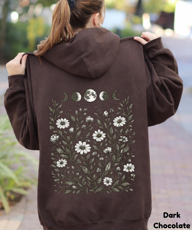 May include: Dark chocolate brown hoodie with a floral and moon phase design on the back. The graphic features white daisies, green leaves, and a series of moon phases. The text "Dark Chocolate" is at the bottom right.